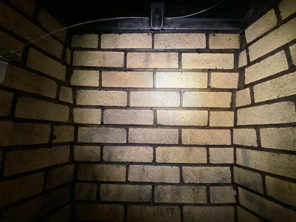 A dimly lit corner of a brick wall with tan bricks and dark mortar hints at a story untold. High above, shadowy structures could be chimney caps from Fairfield County, CT, adding intrigue as a wire weaves along the top edge. The lighting casts a subtle pattern on the bricks.