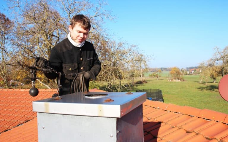 A person stands on a red-tiled roof in New Haven County, CT, holding chimney sweeping equipment—a brush and rope. The open chimney cap reveals a clear blue sky and a rural landscape with trees and fields stretching into the distance.