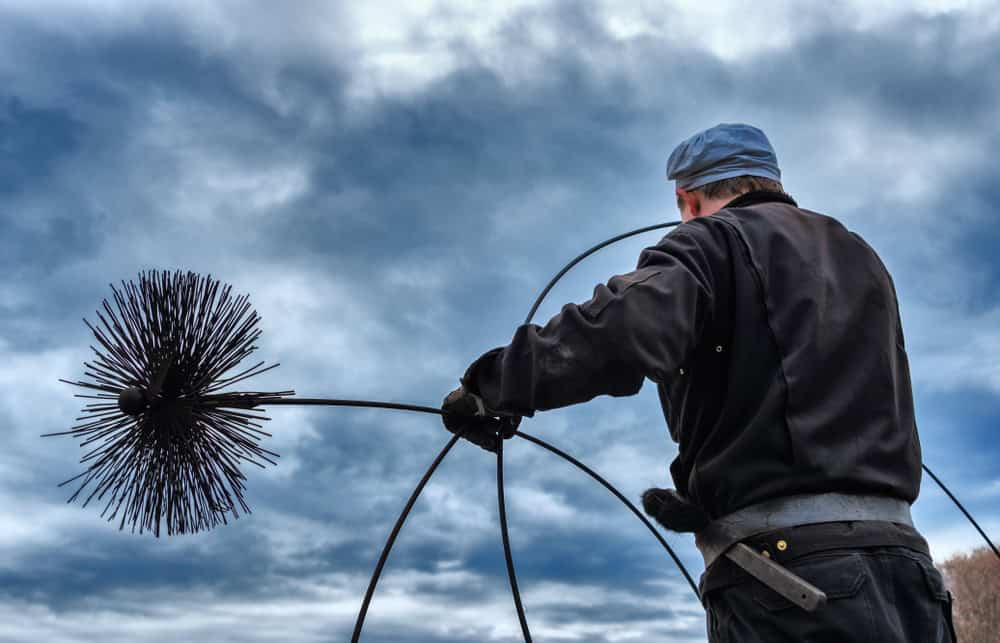 A chimney sweep from New Haven County CT, clad in dark clothing and a blue cap, holds a chimney brush against the cloudy sky. The long-bristled brush, essential for cleaning chimneys, reflects his dedication to maintaining safe fireplaces.