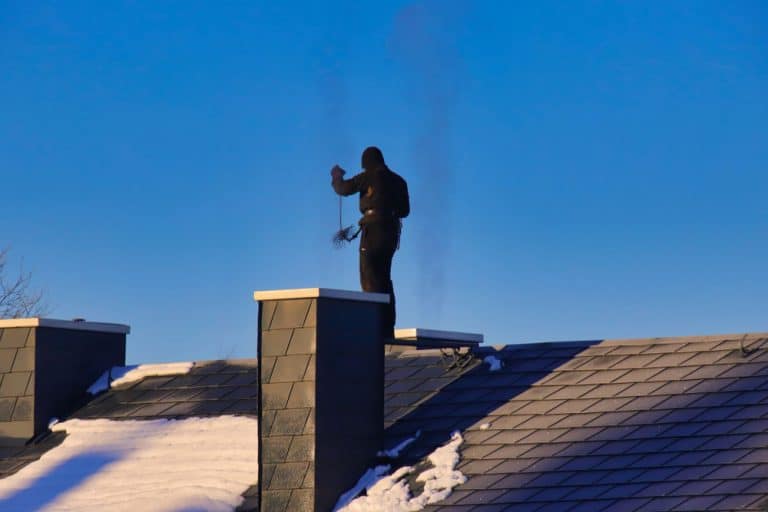 A person dressed in black stands atop a snowy roof, silhouetted against a clear blue sky, expertly wielding chimney cleaning tools near two chimneys, ready for a chimney sweep in New Haven County CT.