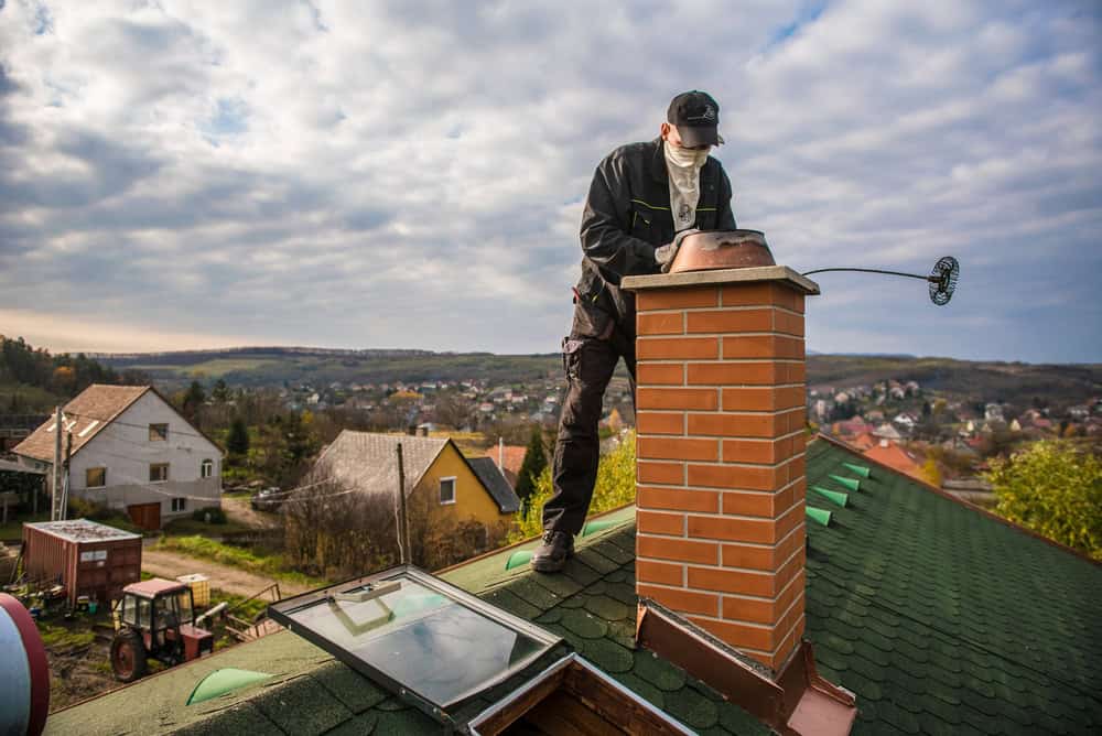 A worker in protective gear stands on a green shingled roof, inspecting a red brick chimney with a chimney sweep brush. Perhaps it's time for some chimney repair in Fairfield County, CT. The background features a rural landscape with scattered houses and cloudy skies.