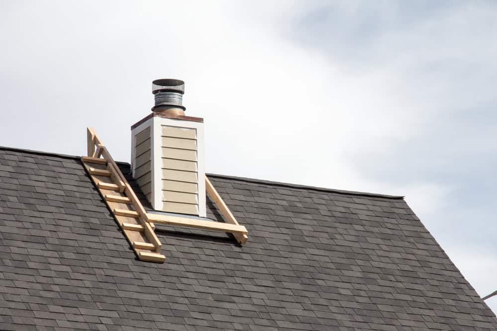 A wooden ladder leads to a small chimney on a shingled roof, perfect for any needed chimney repair in Fairfield County, CT. The sky is partly cloudy.
