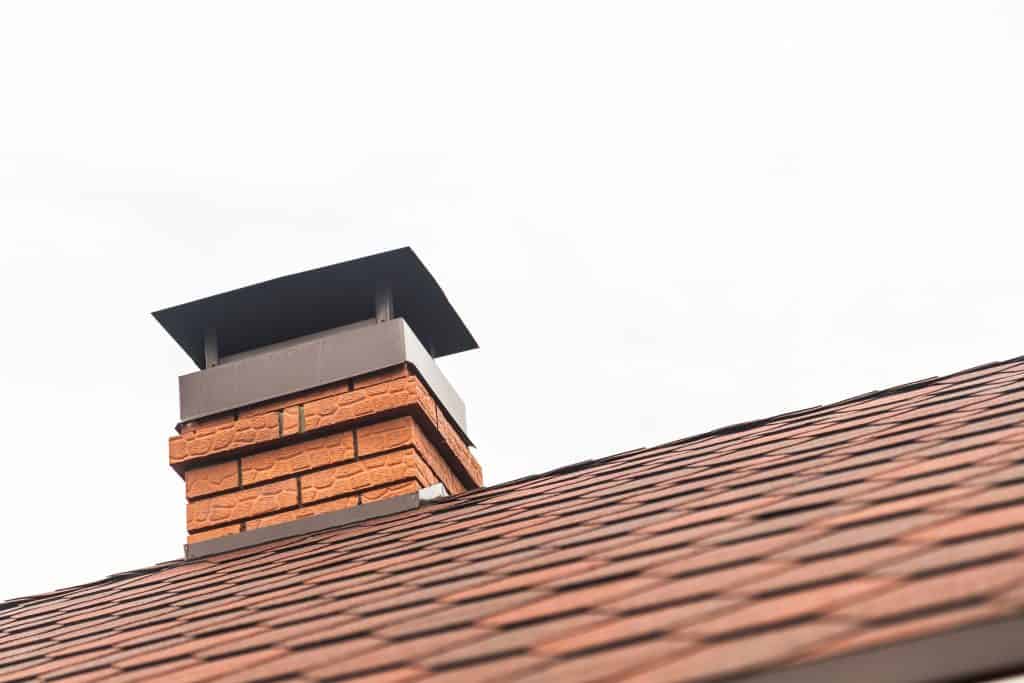 Close-up of a brick chimney with a metal cap, showcasing expert Chimney Caps Fairfield County CT craftsmanship, on a house roof covered in brown shingles, set against an overcast sky.