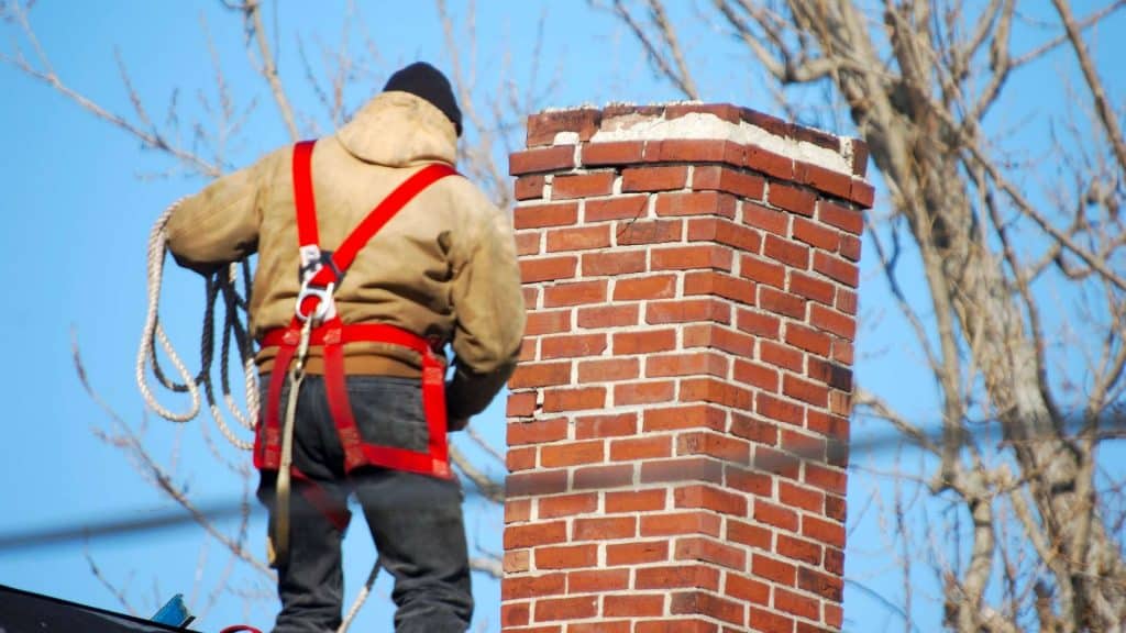 A person wearing a tan jacket, black hat, and red safety harness stands on a roof next to a brick chimney. Holding a rope for chimney repair in Fairfield County CT, they work efficiently under clear blue skies and bare trees.