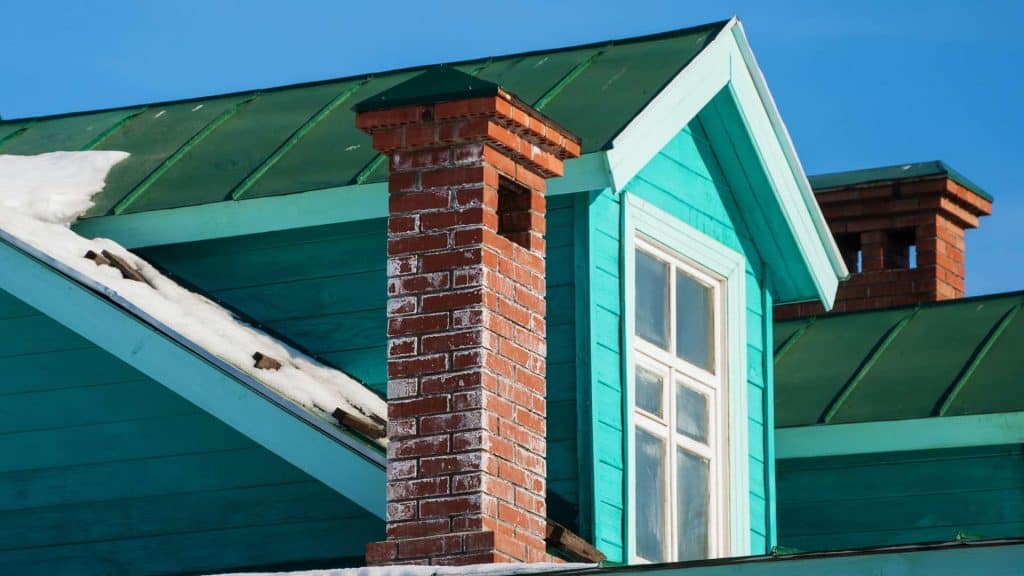 A vibrant turquoise house roof with a snow-capped section and a prominent red brick chimney stands out beautifully. The clear blue sky forms the backdrop, highlighting the house's rich colors. Nearby, Chimney Caps Fairfield County CT ensures every chimney is protected amidst nature’s elements.
