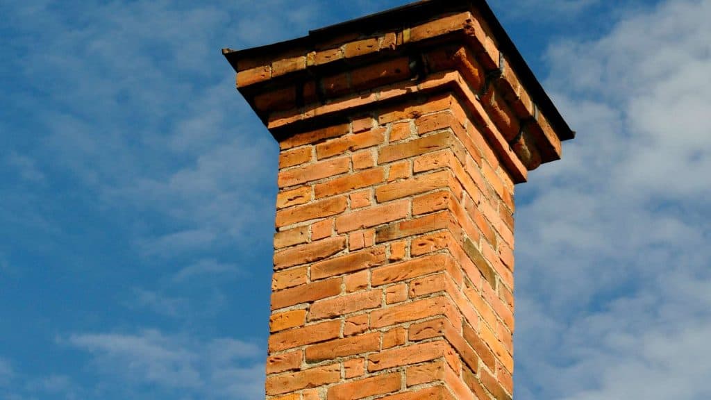 A tall brick chimney against a blue sky with scattered white clouds stands proudly, its rectangular shape and dark flat top hinting at times past. The orange, weathered bricks offer a rustic charm. Consider expert Chimney Repair Fairfield County CT to maintain its enduring beauty.