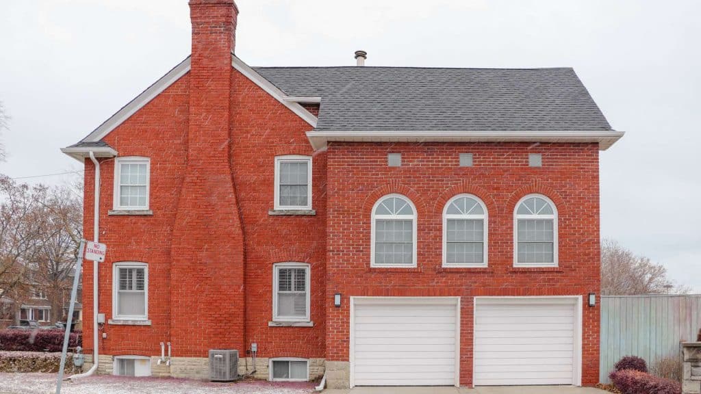 A red brick house with a black shingled roof and a prominent chimney, perfect for those considering chimney repair in Fairfield County, CT. The building has symmetrical windows and two white garage doors. A small "no parking" sign is visible on the left, set against an overcast sky.