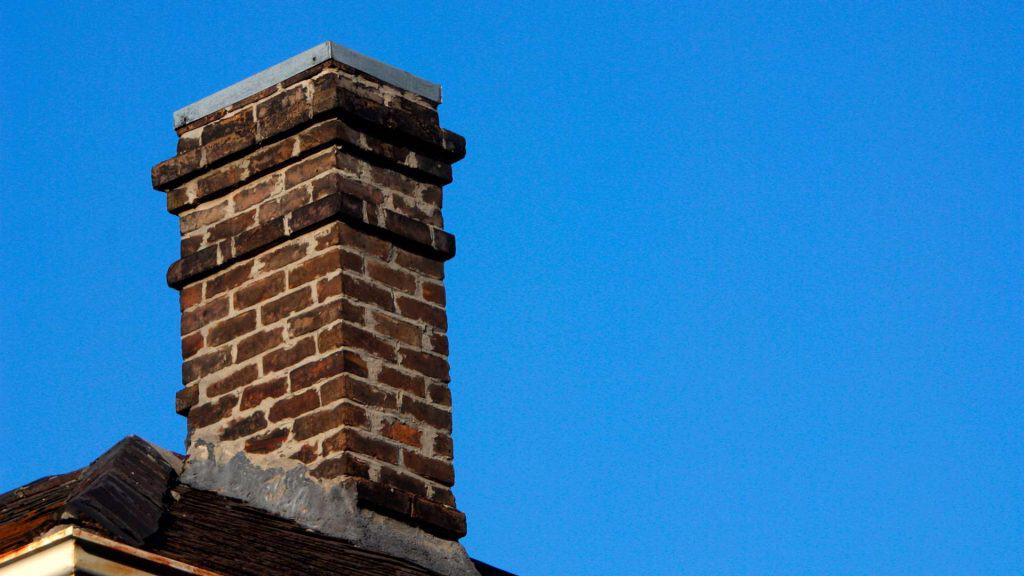 A brick chimney extends upward from a rooftop against a clear blue sky, showcasing its reddish-brown bricks and grey cap. Recently serviced by Chimney Sweep New Haven County CT, the structure stands sturdy. Part of the roof with dark shingles is visible in the lower left corner.