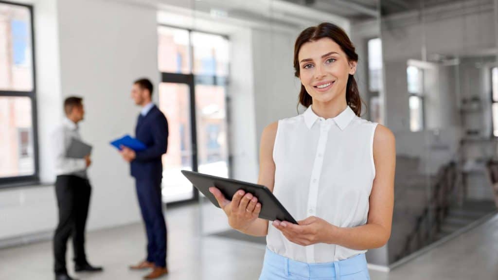 A woman in a sleeveless blouse holds a tablet and smiles in a modern office. Sunlight pours through large windows, brightening the space where two men in suits chat in the blurred background. Just as chimney repair Fairfield County CT professionals ensure safety, this team focuses on innovation.