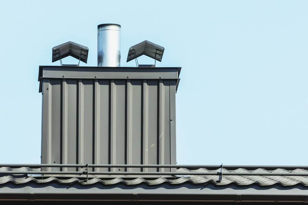 A rooftop with a modern metal chimney featuring two vents and a central exhaust pipe gleams under the clear blue sky. The roof, adorned with corrugated metal sheets, might soon benefit from quality Chimney Caps in Fairfield County CT to ensure lasting durability.