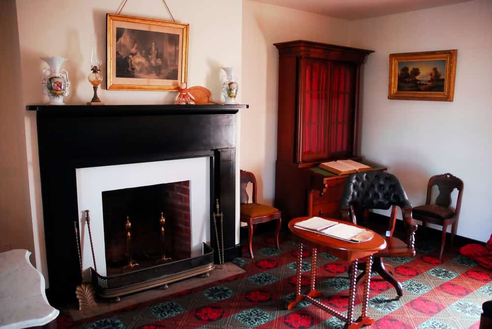 A cozy room with antique furniture, featuring a black fireplace fitted with chimney caps from Fairfield County, CT. There’s a wooden cabinet and a small table with books. Two chairs are placed around the fireplace, and artwork adorns the walls. The carpet boasts a red and green pattern.