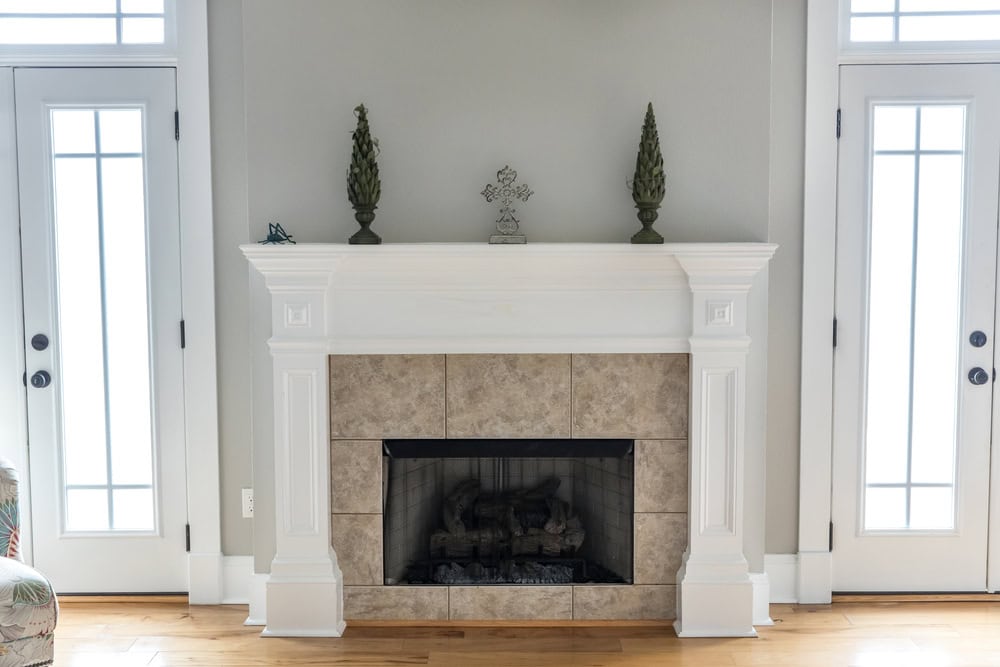 A simple, white wooden fireplace mantel with decorative columns on each side complements the room. Two small topiary sculptures sit proudly on the mantel, while glass doors flank the fireplace, inviting natural light in. Consider adding chimney caps in Fairfield County, CT for added safety and style.