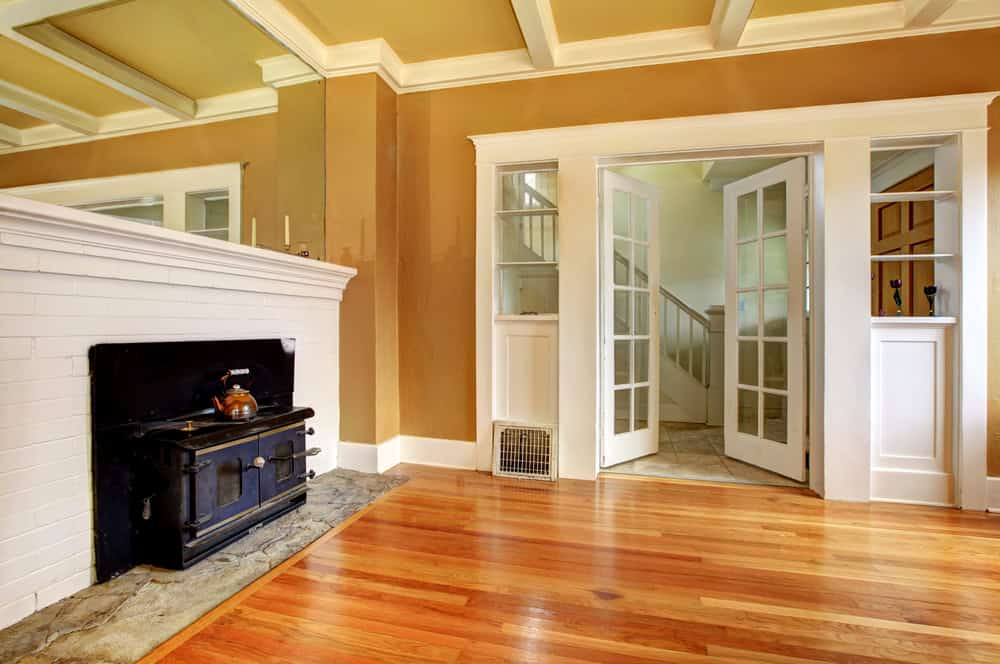 A cozy living room features warm wooden flooring and a white brick fireplace with a sleek black stove insert. Mustard-colored walls complement the large mirror above the fireplace, while French doors lead to a hallway with visible stairs, showcasing recent chimney repair from Fairfield County, CT.