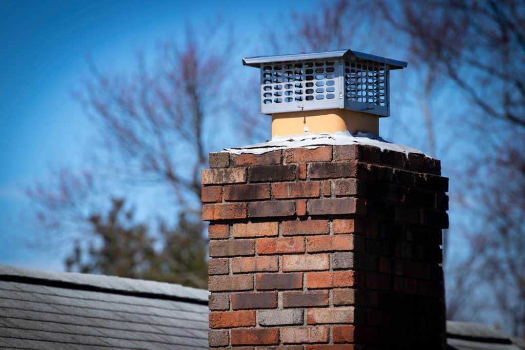 A close-up of a brick chimney with a metal cap in Fairfield County CT, set against a clear blue sky. This chimney, ready for any repairs needed, is part of a building roof, with snow dusting the top bricks. Bare tree branches elegantly frame the background.