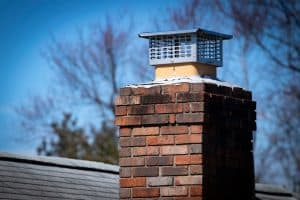 A close-up of a brick chimney with a metal cap in Fairfield County CT, set against a clear blue sky. This chimney, ready for any repairs needed, is part of a building roof, with snow dusting the top bricks. Bare tree branches elegantly frame the background.