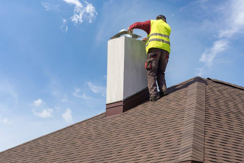 A worker in a bright yellow safety vest stands on a shingled rooftop, inspecting a chimney for potential repair in Fairfield County, CT. The sky is clear with a few clouds floating by.
