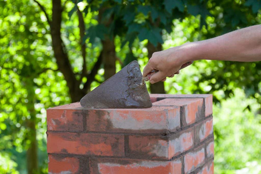 A person is using a trowel to apply mortar to the top of a red brick column, reminiscent of chimney repair work in Fairfield County, CT. Lush green trees are blurred in the background, suggesting an outdoor setting.