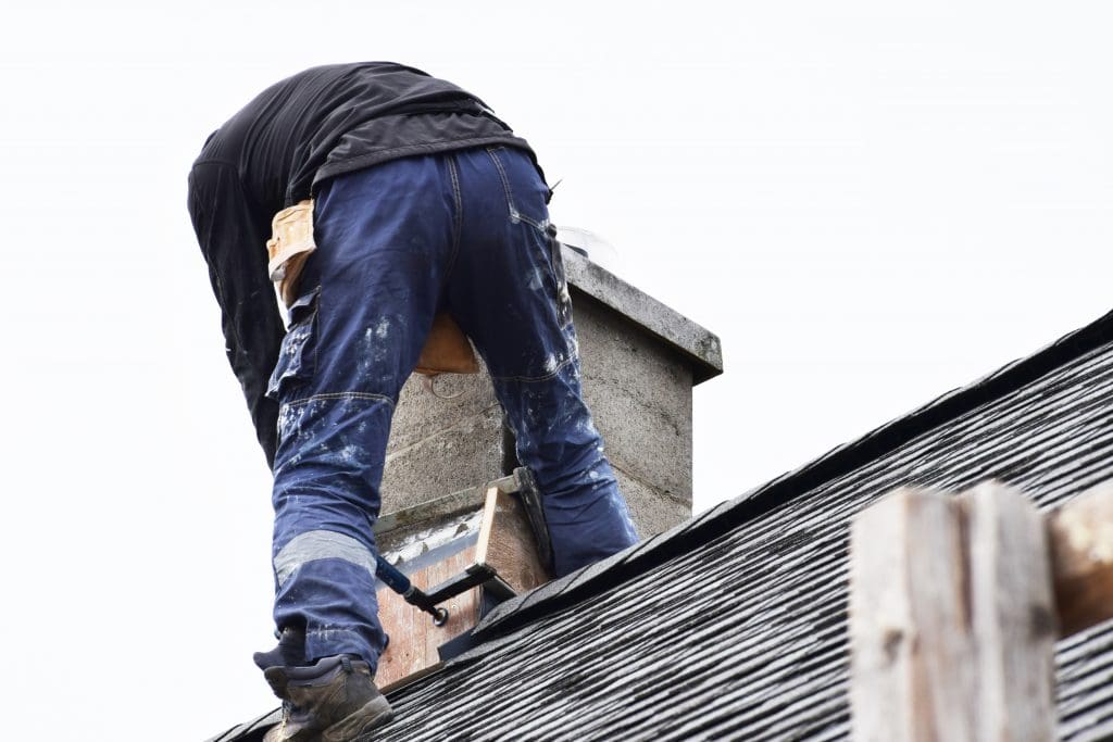 A worker, seen from behind, is expertly repairing a chimney on a rooftop in Fairfield County, CT. They are wearing a black jacket and blue work pants with paint stains, standing on dark roof shingles. The overcast sky looms above as they focus on the chimney repair.