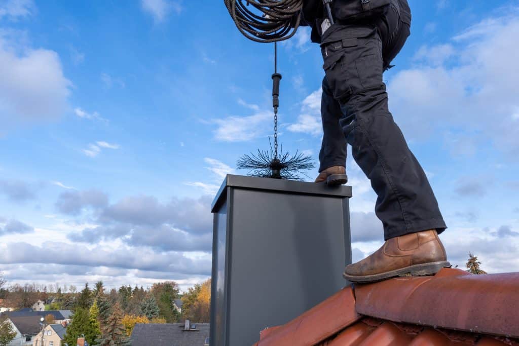A person stands on a rooftop in New Haven County, wearing brown boots, holding a chimney cleaning tool attached to a cable. The chimney features a brush at the end of the tool. The background shows a cloudy sky and neighborhood houses. It's another day for the dedicated chimney sweep.