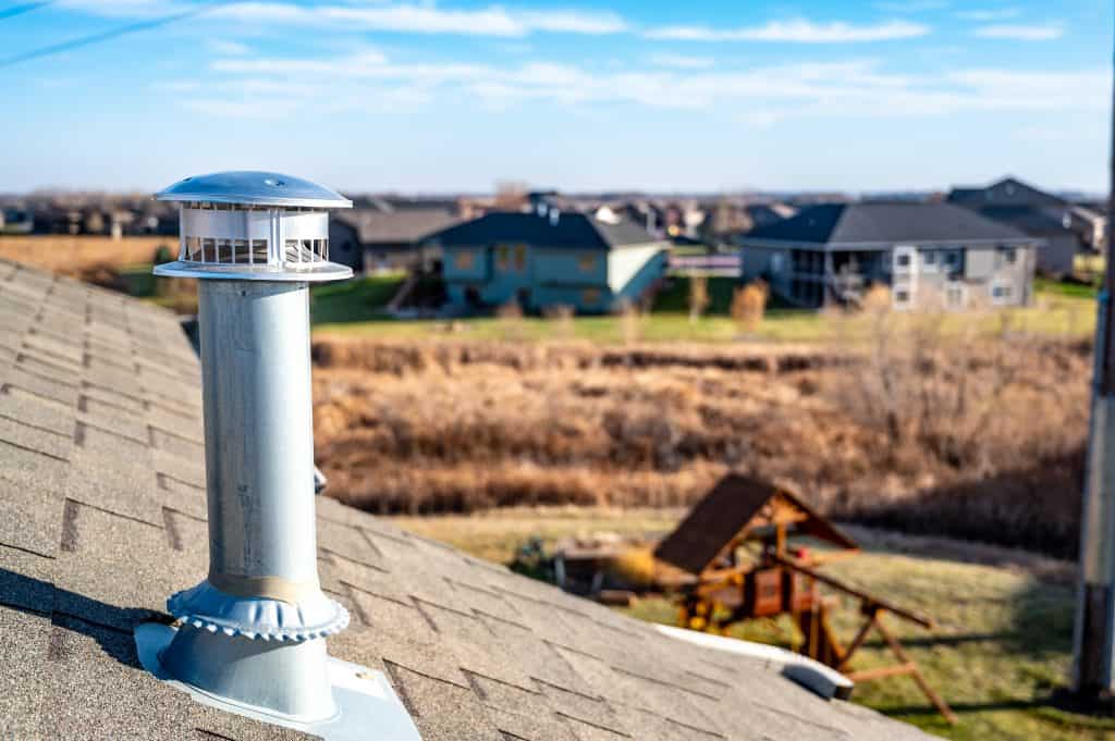 A metal chimney vent, complemented by a sturdy chimney cap, is installed on a shingled roof. It overlooks a suburban neighborhood with houses and open fields. The sky is clear and sunny, perfect for anyone considering chimney repair in Fairfield County, CT.