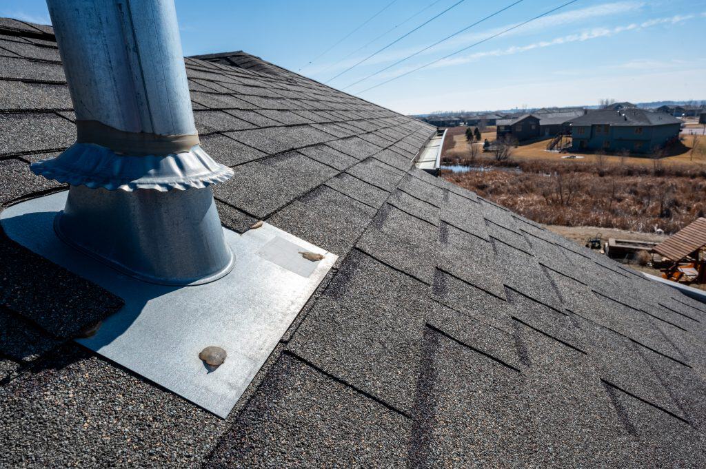 A sloped roof with gray asphalt shingles and a metal vent pipe is visible in the foreground, alongside a newly installed chimney cap. The background features a suburban landscape with houses, grassy fields, and a clear blue sky.