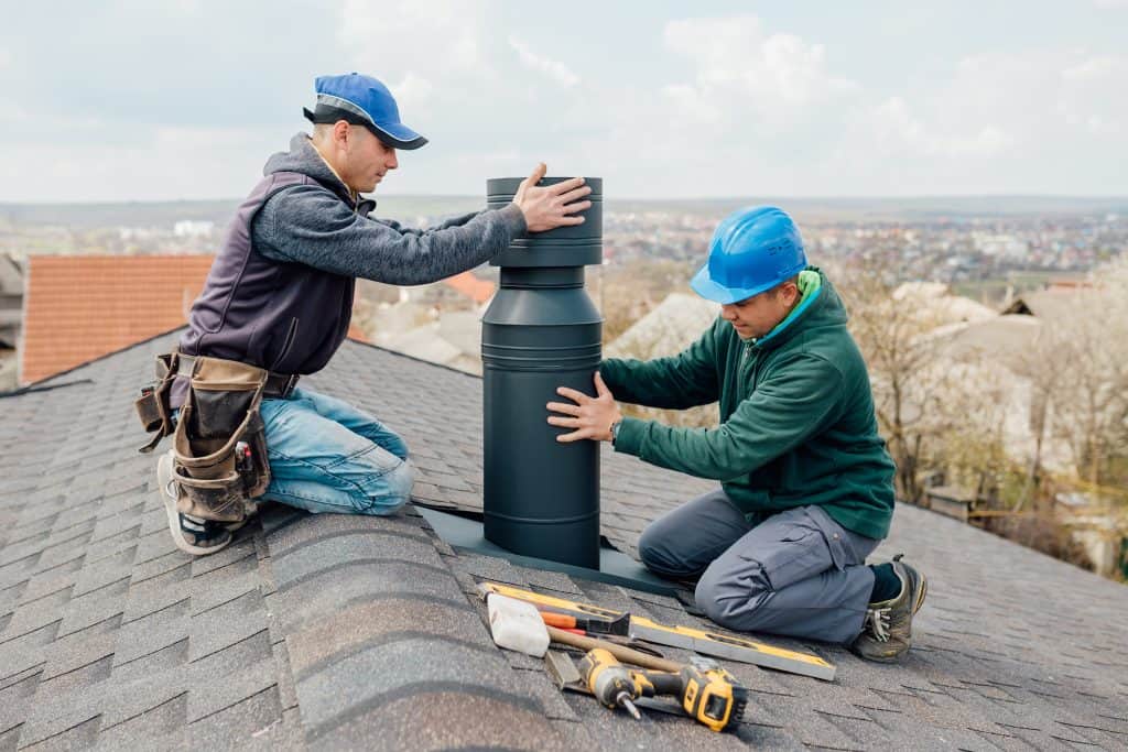 Two workers in blue hats are installing a chimney cap on a shingled roof. Various tools, including a drill and measuring tape, are scattered nearby. The background shows a clear sky and a view of rooftops in the distance, resembling scenes from Chimney Repair Fairfield County CT projects.
