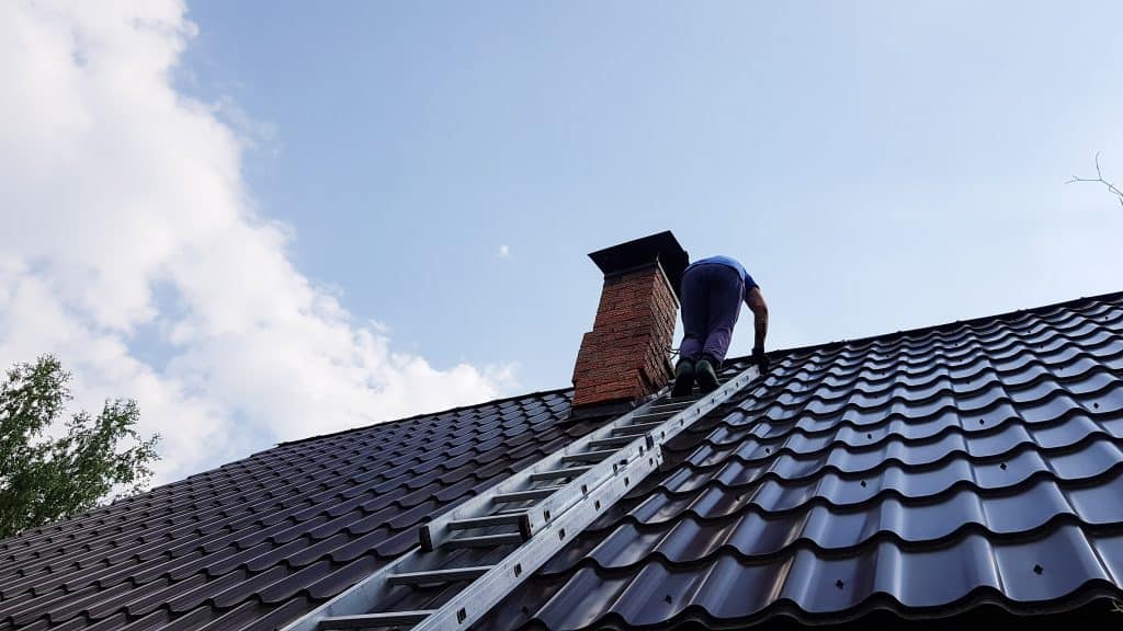 A person climbs a metal ladder onto a roof near a chimney under a partly cloudy sky, ready for chimney sweep services in New Haven County, CT.