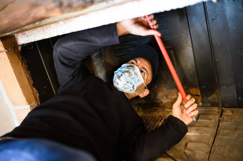 A person wearing a face mask and cap lies on their back under a fireplace, using a red tool to clean or repair it. This scene, set in Fairfield County CT, captures an indoor maintenance activity involving chimney caps.
