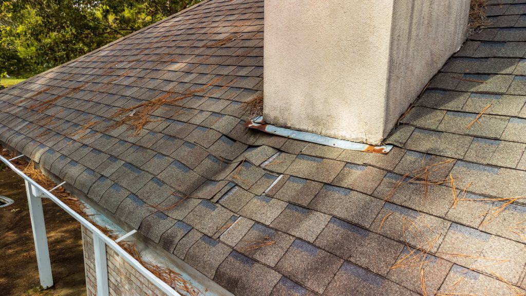 A sloped roof with grey shingles showing signs of damage and warping, possibly indicating the need for chimney repair in Fairfield County CT. Pine needles are scattered across the surface, and a chimney is visible. The roof's edge is lined with a gutter.