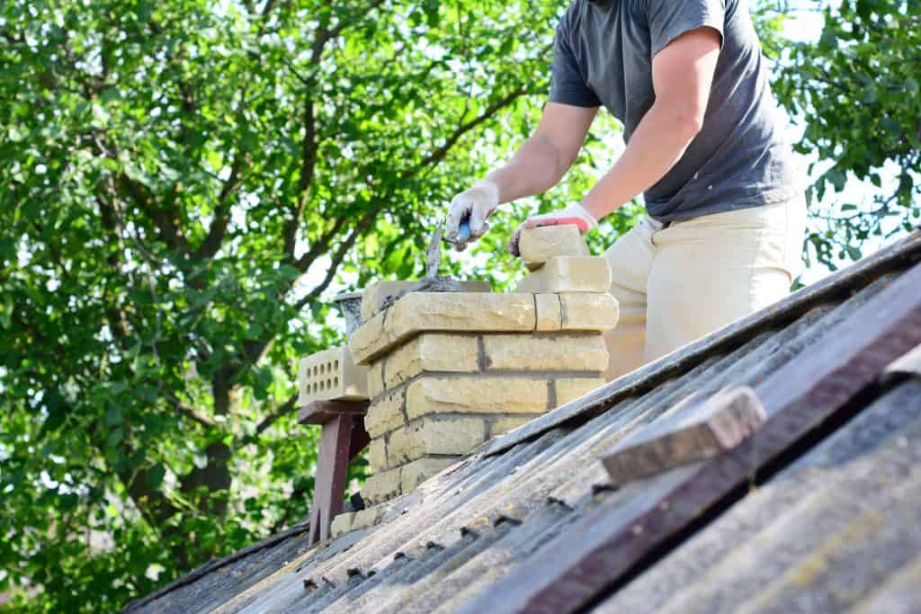 A person is working on chimney repair in Fairfield County, CT, expertly placing bricks on a sloped roof. Wearing gloves for safety, they focus intently as a tree with green leaves sways gently in the sunny background.