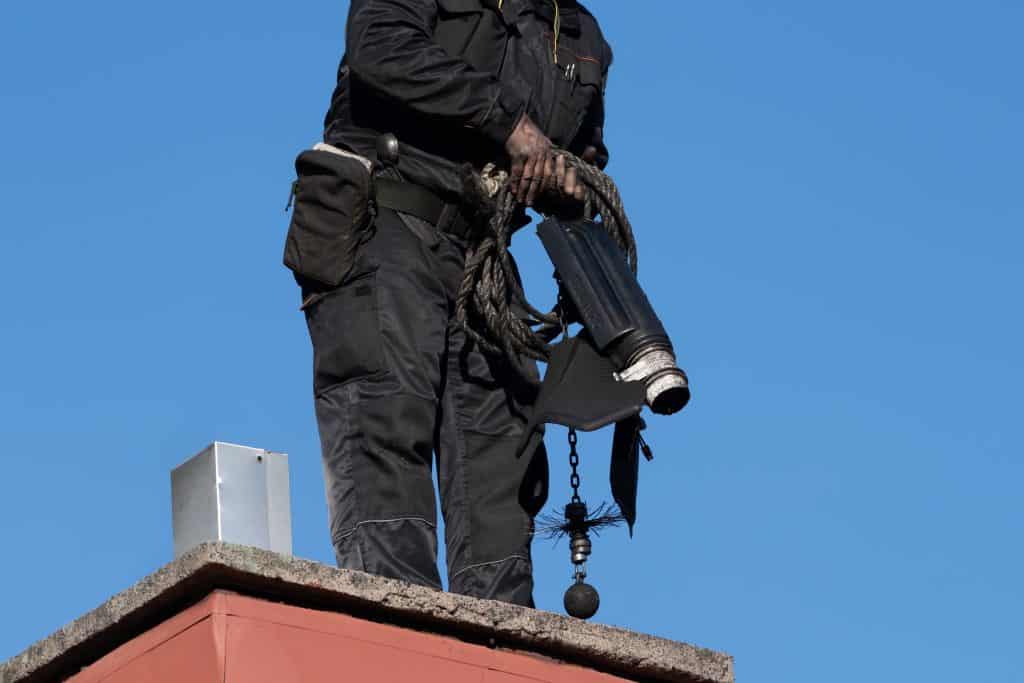 A person in a black uniform stands on a rooftop, holding chimney cleaning equipment, including a sweeper brush and rope. The clear blue sky serves as the backdrop, as they expertly perform their duties for Chimney Sweep New Haven County CT.