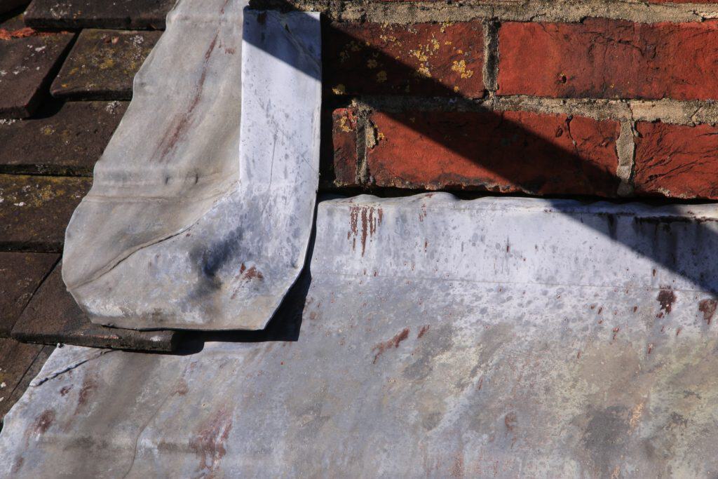 Close-up of a weathered roof showcasing the intersection of metal flashing and red brick, adjacent to a chimney cap. The oxidized metal casts a shadow on the bricks, which have visible mortar and patches of moss—a scene typical for chimney repairs in Fairfield County CT.