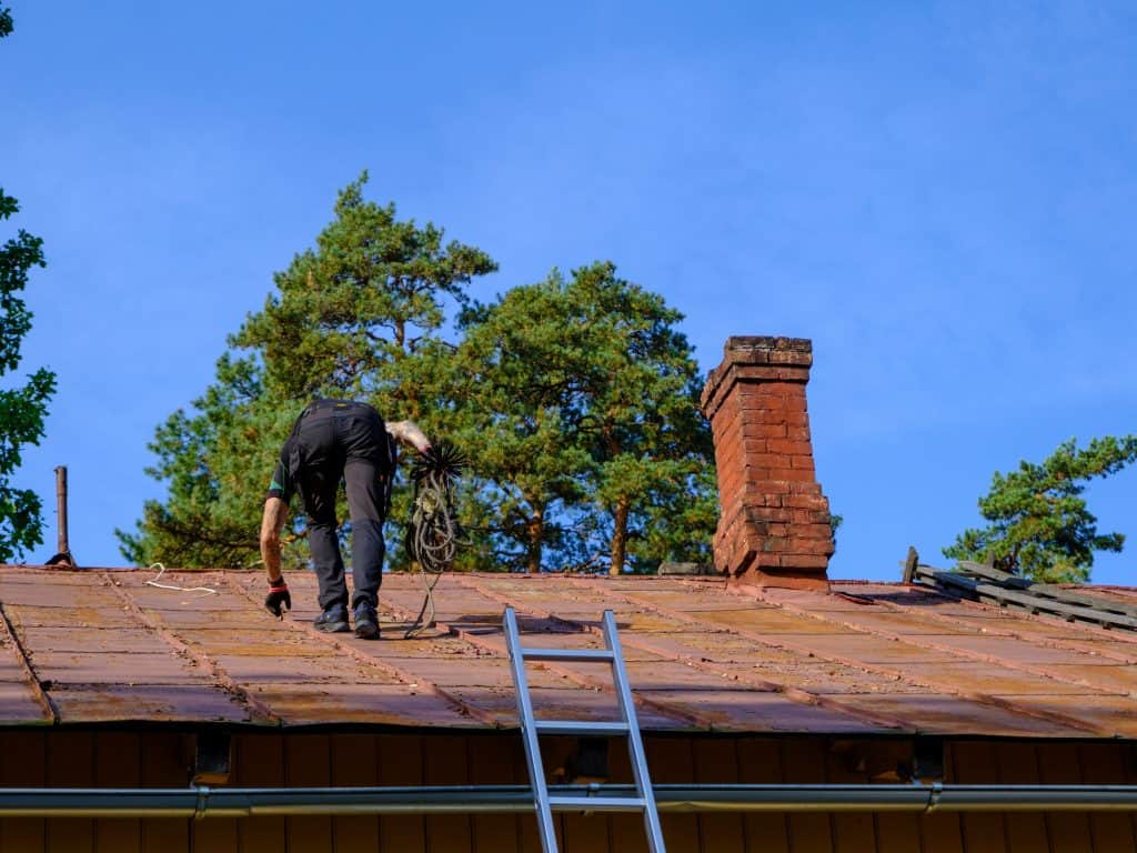 A person repairs the roof near a ladder, focusing on chimney repair in Fairfield County, CT. The brown roof features a red brick chimney, framed by trees and a clear blue sky.