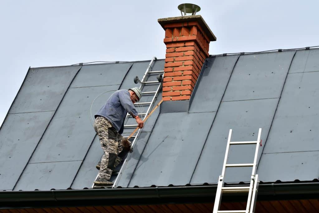A worker on a roof with a ladder leans against a chimney, diligently securing something with a tool. Another ladder is positioned at the roof's edge. The roof appears steep, and the day is clear—a scene reminiscent of a skilled Chimney Repair in Fairfield County, CT.
