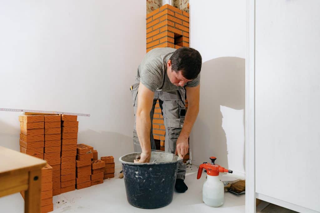 A man wearing a gray t-shirt and work pants bends over a large bucket, mixing cement for a chimney repair in Fairfield County CT. He's in a room with white walls; a stack of bricks is nearby. A red and white spray bottle is on the floor beside him.