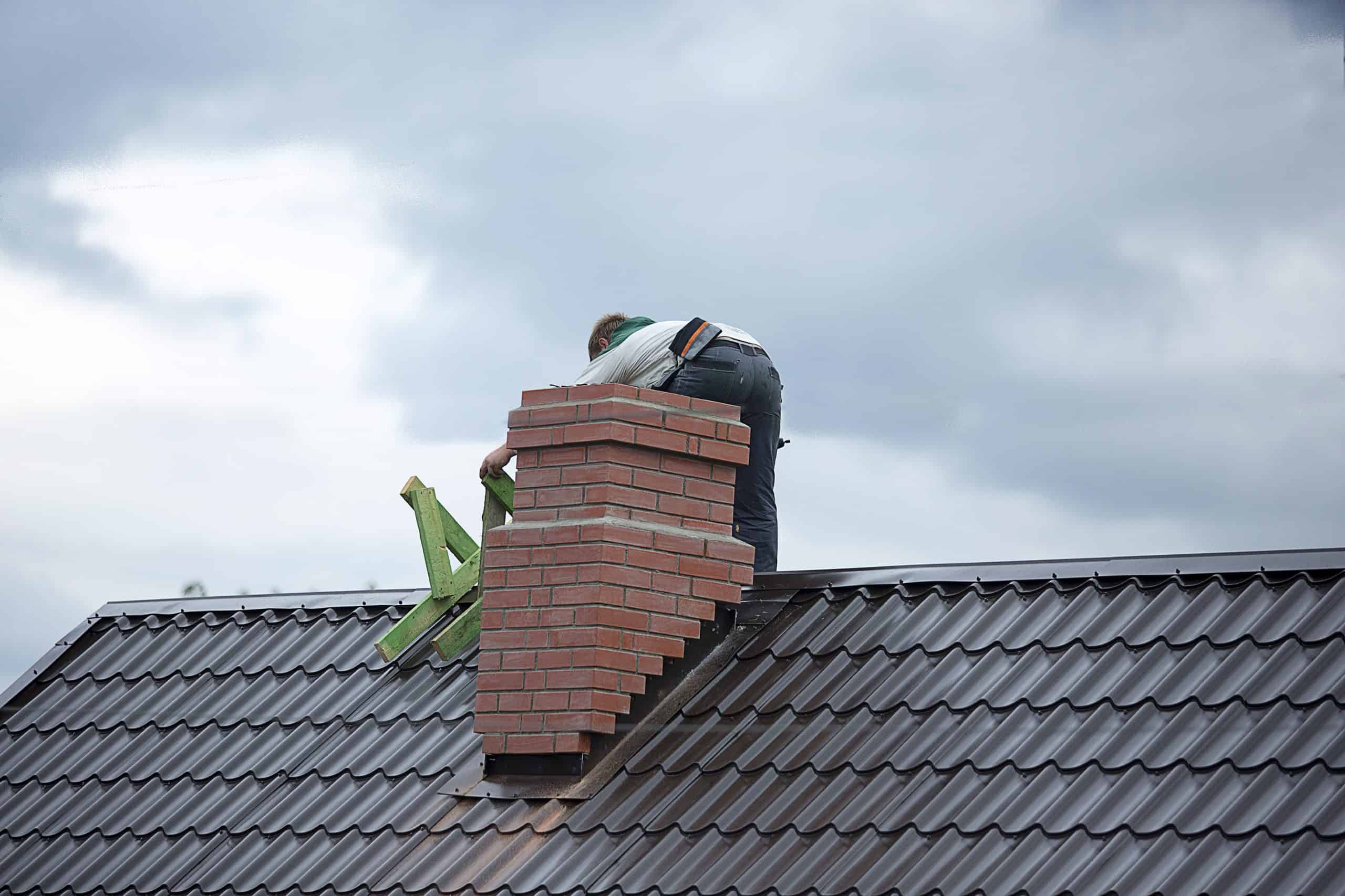 A worker meticulously tends to chimney repairs on a metal roof in Fairfield County, CT, balancing on a green ladder. Above, the sky is overcast with grey clouds, adding a moody backdrop to the task at hand.