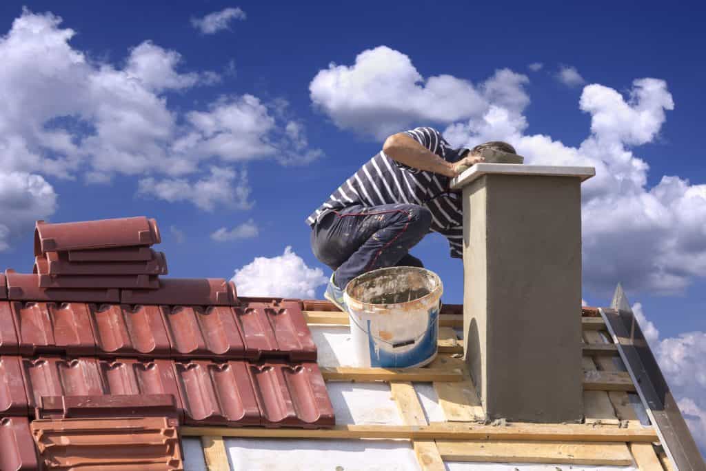 Under a blue sky dotted with clouds, a person busily works on chimney repair in Fairfield County, CT. Surrounded by red roof tiles, they carefully apply material to the chimney, while a bucket rests nearby on the partially constructed roof.