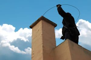 A chimney sweep stands on a rooftop in New Haven County, CT, holding a wire tool above a chimney. Against the backdrop of blue sky and fluffy white clouds, the silhouette wears dark clothing and a harness, ensuring safety and precision.