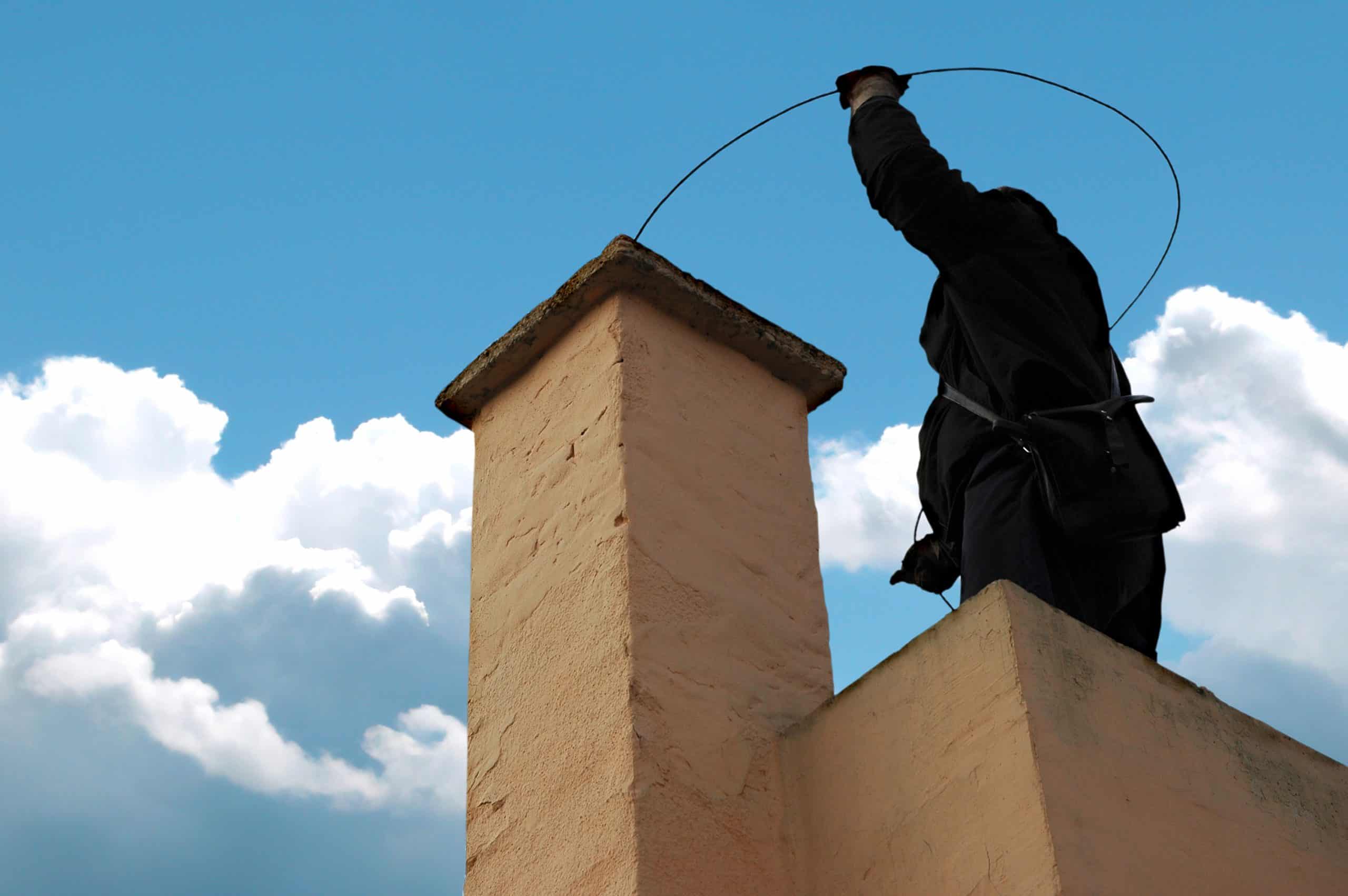 A chimney sweep stands on a rooftop in New Haven County, CT, holding a wire tool above a chimney. Against the backdrop of blue sky and fluffy white clouds, the silhouette wears dark clothing and a harness, ensuring safety and precision.