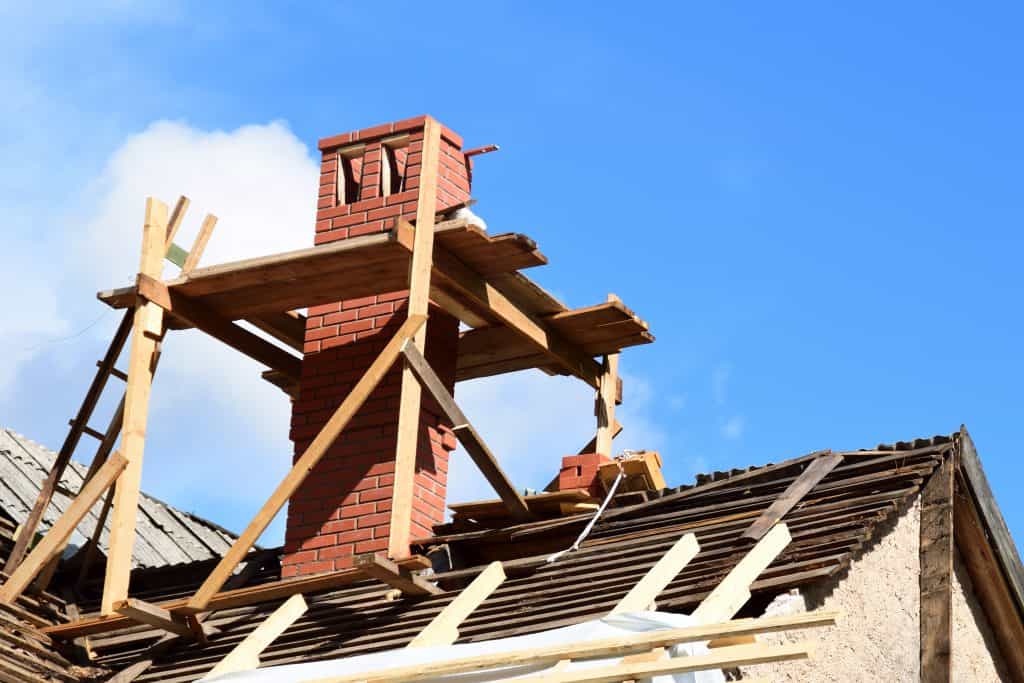 A red brick chimney is under construction, supported by a wooden scaffolding on a rooftop. The roof appears partially dismantled, with some exposed beams and roofing material. Skilled in chimney repair in Fairfield County, CT, the crew works efficiently under the clear blue sky.